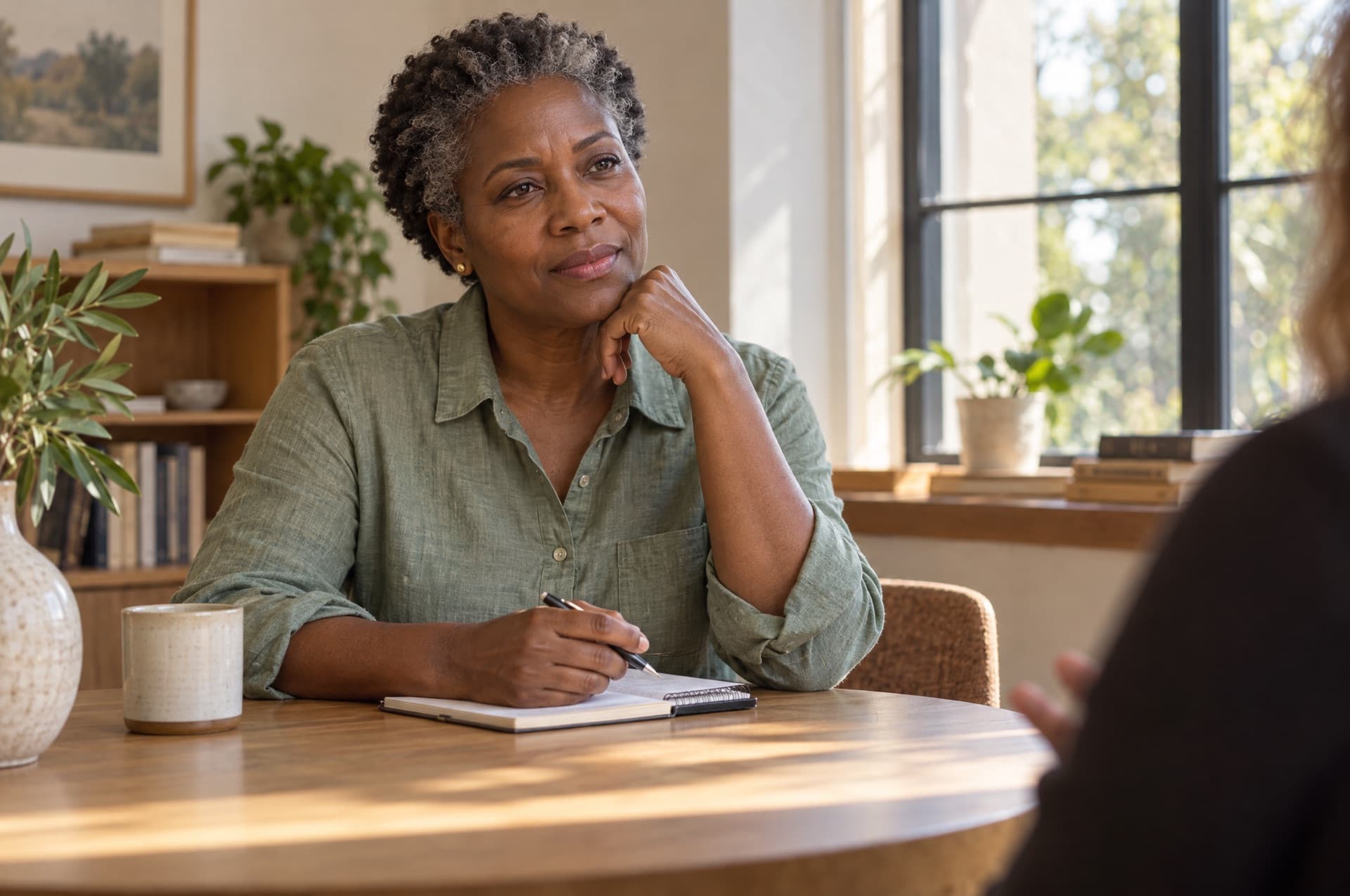 A woman in her early fifties with deep brown skin and short natural gray-streaked hair sitting at a table in a calm, sunlit room, listening and holding a notebook