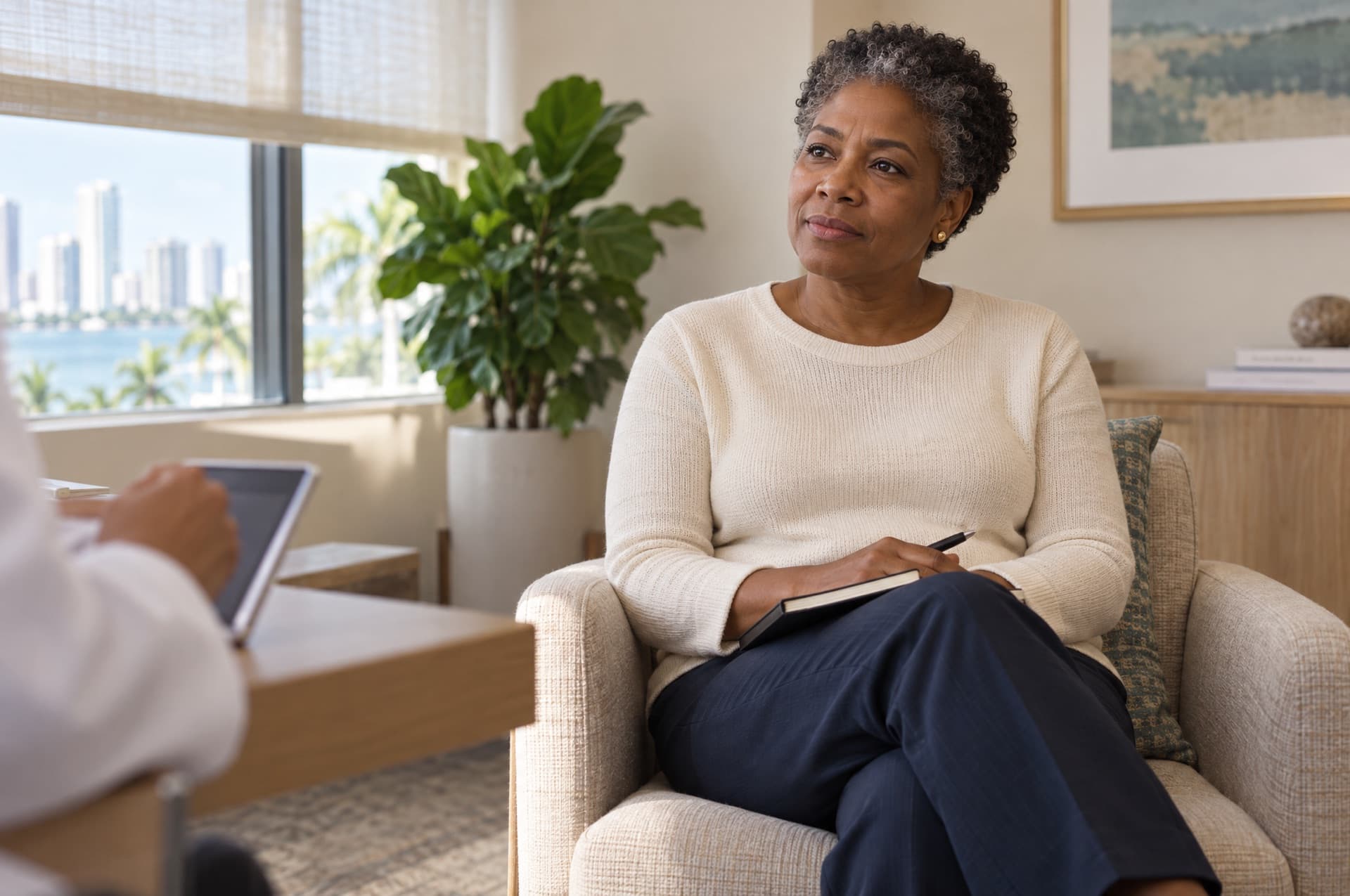 A woman in her early fifties with short natural gray-streaked hair sits in a calm consultation room, listening and holding a notebook on her lap
